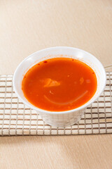 A white bowl of tomato-based vegetable soup placed on a beige mat, showing bright red broth.