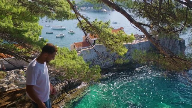 Close-up of man in white shirt sitting on cliff with stunning view of green hills and bright blue Adriatic Sea Petrovac Riviera coast