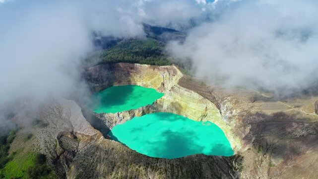 Drone aerial of Kelimutu volcano featuring its iconic turquoise crater lakes surrounded by dramatic volcanic terrain and drifting clouds in Flores, Indonesia.