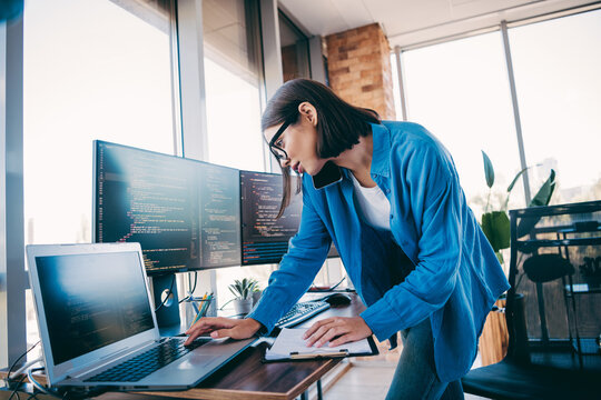 Female developer in a blue shirt works at a modern loft office with a laptop and multiple monitors while coding and planning projects