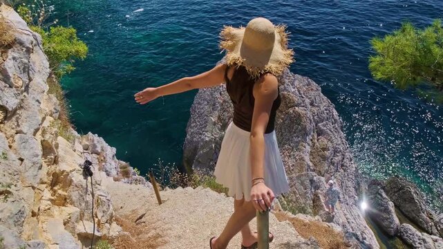 Back view of girl in straw hat raising arms and flowing white skirt standing on narrow cliff path above bright blue Adriatic Sea Petrovac Montenegro