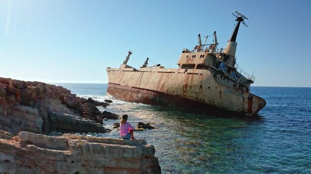 Woman sits on jagged rocks along the Mediterranean coast, gazing at the rusted Edro III shipwreck half-submerged in turquoise water under a clear blue sky in Pegeia, Cyprus