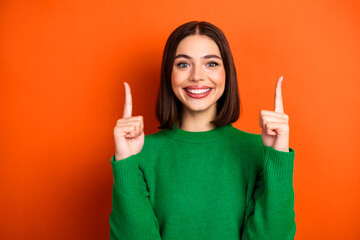 Happy young woman in a green sweater points up with both fingers against an orange background for a...