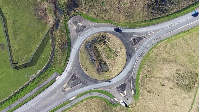 Unfinished roundabout from above with two incomplete junctions as two cars drive across. Drone top-down traffic movement, roadworks, infrastructure, countryside roads.