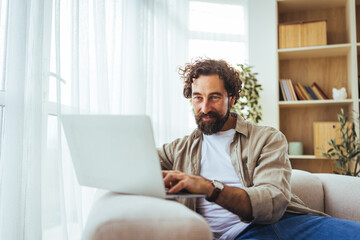 Bearded Man Working Remotely On Laptop At Home Sitting On Sofa In Casual Outfit