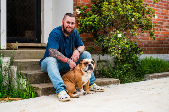 Man and bulldog sit together on concrete steps in front of a brick house with plants