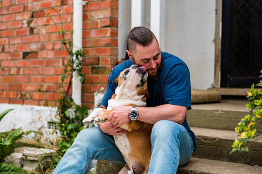 A man sitting on a concrete step holding a dog licking his face