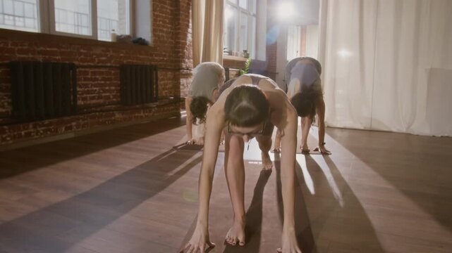Medium full tracking shot of young Caucasian female hatha instructor leading yoga class for women in sunlit loft studio, demonstrating sun salutation sequence and explaining asana technique
