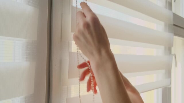 Close up shot of a woman's hand opening white roller blinds on a window.
