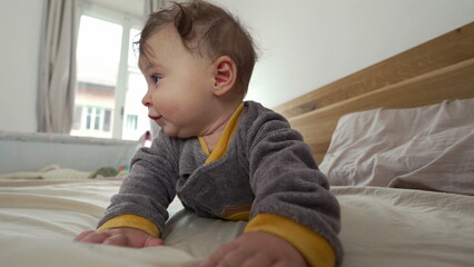 Baby lying on stomach on bed, propping up with arms and looking to the side with curious expression, early motor skill development, cozy indoor family environment, exploration and growth © Marco