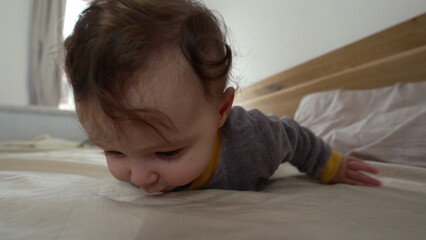 Baby lying on stomach on bed, propping up with arms and looking to the side with curious expression, early motor skill development, cozy indoor family environment, exploration and growth © Marco