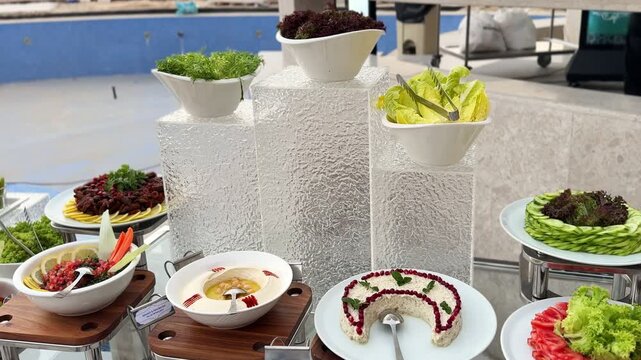 Upscale outdoor salad bar featuring hummus, mixed greens, sliced tomatoes, cucumbers, tabbouleh, and artfully arranged vegetable platters on modern stands near poolside.