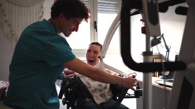 Young male caregiver assisting a laughing disabled woman in a wheelchair, using a patient hoist lift for safe transfer and mobility inside a house, demonstrating compassionate home healthcare