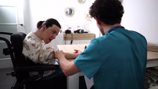 Young male caregiver providing home assistance and physical therapy to a woman with cerebral palsy, helping with her arm movements and exercises at a table in a bright, modern living room