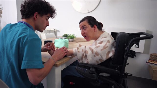 Supportive male nurse providing home care to a smiling woman with a disability sitting in an electric wheelchair, holding her hand while they talk and share a positive and tender moment