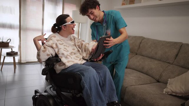 Male nurse assisting a cheerful young woman with cerebral palsy sitting in a motorized wheelchair, showing her information on a digital tablet during a home care visit in a modern apartment