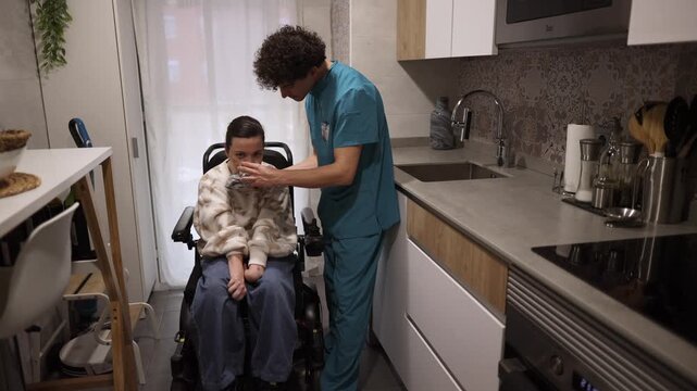 Compassionate caregiver assisting a woman with a physical disability in a wheelchair, helping her drink a glass of water in a modern kitchen, showing support, empathy, and professional healthcare