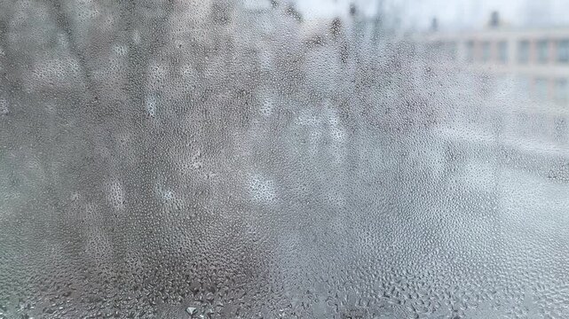 Condensation on window glass as snow falls outside. Water droplets cover window, blurring view of snowy landscape. Winter season with cold weather, frosty window, snowy atmosphere, indoor scene