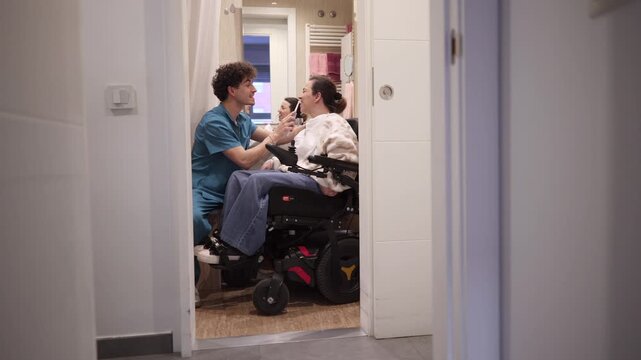 Young male caregiver gently brushes teeth of a smiling woman in a wheelchair during morning bathroom routine, providing compassionate in home personal care and support