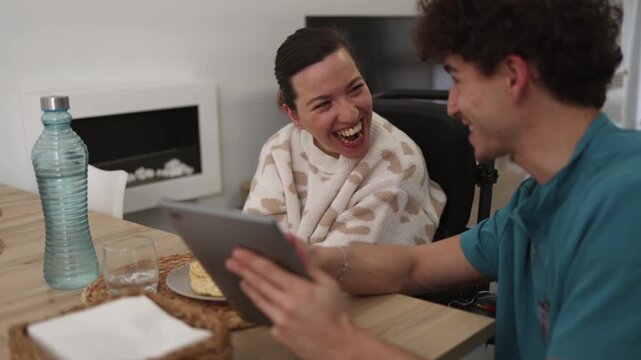 Cheerful disabled woman in a wheelchair laughing happily while a young male nurse shows her something funny on a digital tablet, sharing a moment of joy and connection at home