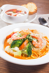 A white plate of spicy Thai noodle soup topped with shrimp, broccoli, carrots, and fresh herbs, served with soup and bread on the side.