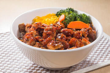 A white bowl filled with diced chicken coated in spicy sauce, served with rice and vegetables like broccoli and carrot.