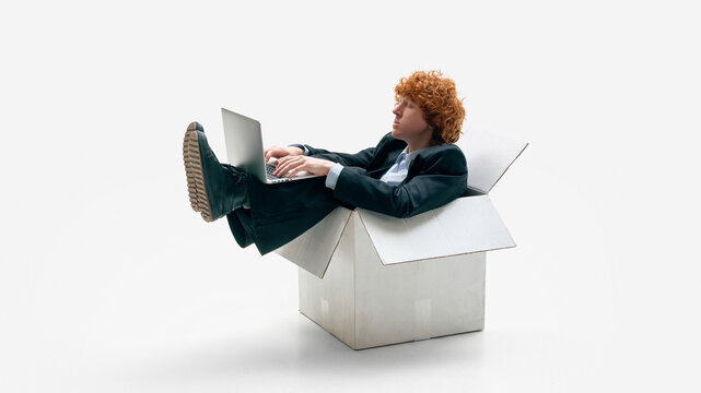 Businessman sitting inside cardboard box working on laptop on white studio background. Concept of startup minimalism, limited resources, bootstrapping and flexible workspace culture.