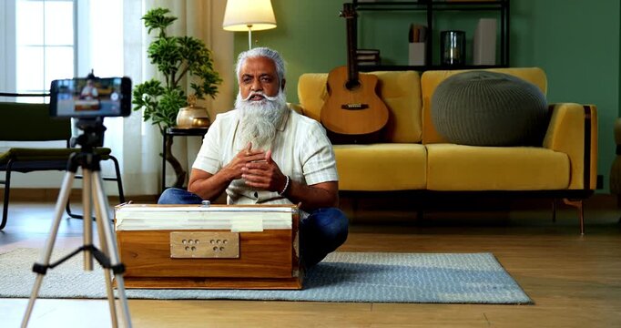 Indian mid age bearded man recording harmonium playing session on smartphone for online class at home, stylish mature music tutor creating classical lesson video with tripod setup in modern studio