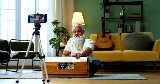 Indian mid age bearded man recording vocals music session on smartphone for online class at home, stylish mature tutor teaching Indian classical singing with harmonium in modern studio