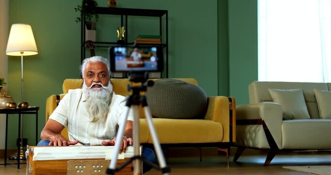 Indian mid age bearded man recording vocals music session on smartphone for online class at home, stylish mature tutor teaching Indian classical singing with harmonium in modern studio