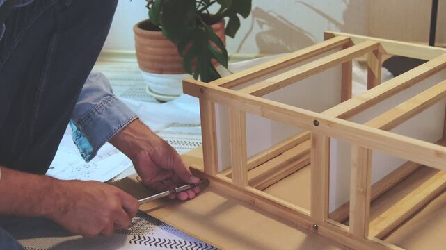 Unrecognizable man assembling a new piece of wooden furniture on the floor with a screwdriver