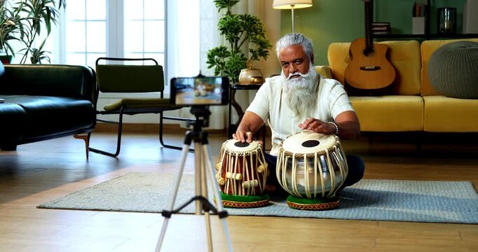 Indian mid age stylish bearded man taking online Tabla musical instrument playing class at home, experienced classical percussion tutor streaming lesson on smartphone in modern studio