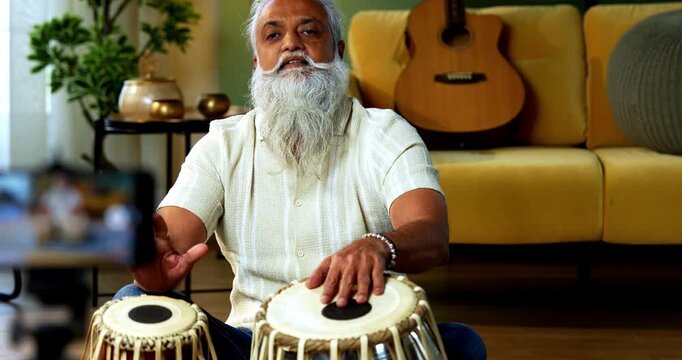 Indian mid age stylish bearded man taking online Tabla musical instrument playing class at home, experienced classical percussion tutor streaming lesson on smartphone in modern studio