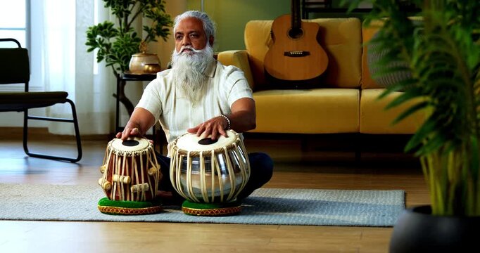 Indian mid age stylish bearded man practicing playing Tabla musical instrument at home, dedicated classical percussion performer doing daily riyaz session in modern home studio interior
