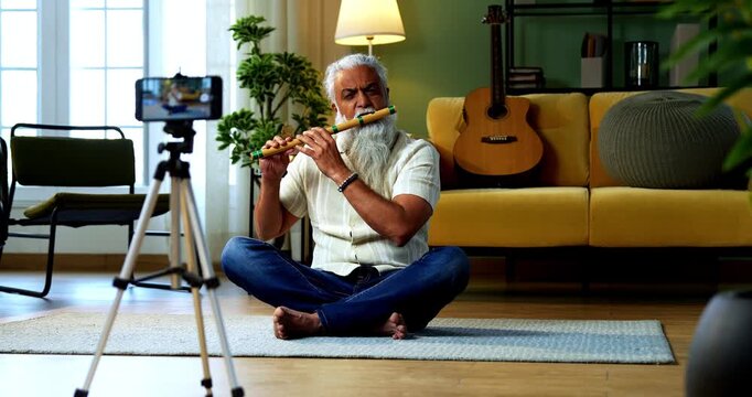 Indian mid age stylish bearded man teaching playing wooden flute musical instrument at home using smartphone, experienced classical bansuri tutor conducting online class in modern studio