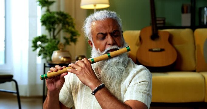 Indian mid age stylish bearded man practicing playing wooden flute musical instrument at home, dedicated classical bansuri performer doing daily riyaz session in modern home studio interior