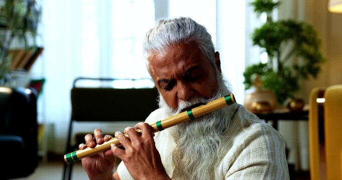 Indian mid age stylish bearded man practicing playing wooden flute musical instrument at home, dedicated classical bansuri performer doing daily riyaz session in modern home studio interior