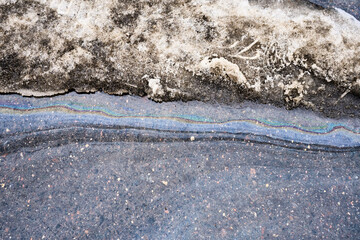 Oil slick creating iridescent rainbow colors on water from melting dirty snow and icy slush, merging onto dark asphalt pavement
