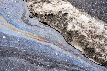 Oil slick creating iridescent rainbow pattern on wet asphalt road next to dirty melting snow