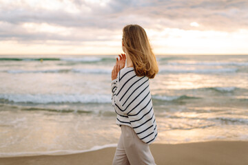 A young woman stands on the beach with her arms crossed. She is looking at the ocean with her eyes. The sun is setting, creating a colorful sky. Waves are gently reaching the shore.