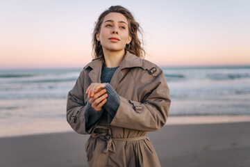 A woman stands on the beach during sunset. She holds her hands to her face as the wind flows through her hair. Waves crash gently in the background while a colorful sky appears. © Koval988