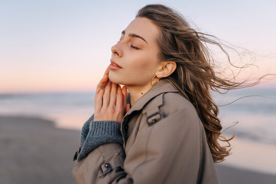 A woman stands on the beach during sunset. She holds her hands to her face as the wind flows through her hair. Waves crash gently in the background while a colorful sky appears.