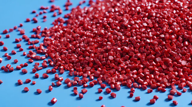 A high-angle close-up shot of bright red plastic granules or polymer pellets scattered on a smooth blue surface, representing raw materials used in industrial manufacturing and recycling processes.