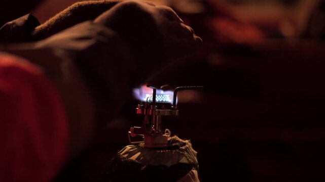 Close-up of a worker warming hands over a portable gas burner with a bright blue flame in a dark, red-lit environment. Selective focus highlights the metal stove and protective gloves, creating a dram