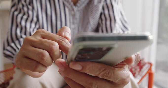Opposite view of unrecognizable Asian woman hands using smartphone on chair by window, scrolling screen with finger in bright home interior, mobile technology and digital lifestyle concept