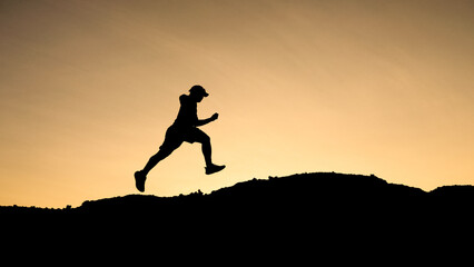 A silhouette of a person jumping in front of a golden hour sky. The person appears to be jumping over a hill. 