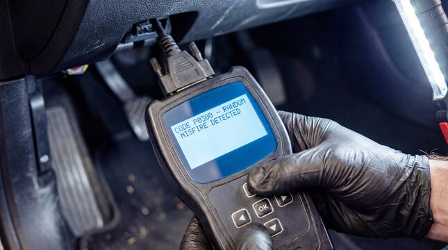 Mechanic wearing black gloves uses an OBD2 scanner displaying diagnostic trouble code P0300 for Random Misfire Detected in a dimly lit car interior.
