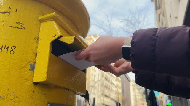 A person drops a letter into a mailbox on the street. The mail is being sent, with cityscapes visible in the background.