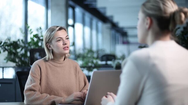 Two women are sitting at a desk with a laptop in front of them