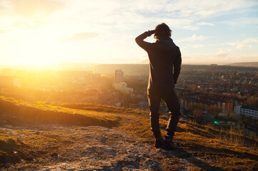 Silhouette of a Female figure standing in a fleece jumpsuit on a hilltop at sunset, overlooking a cityscape with buildings and distant mountains in the background © yanik88